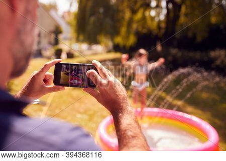 Father Taking Photo Of Daughter Playing In Paddling Pool On Mobile Phone