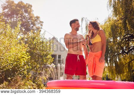 Couple Wearing Swimming Costumes Standing In Paddling Pool In Summer Garden At Home Drinking Beer