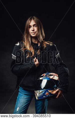 Female Motorcyclist Dressed In Black Jacket With Jeans Poses In Dark Background Holding Protective H