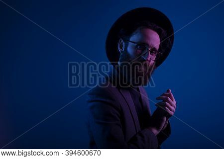 Portrait Of A Bearded Man In Hat And Eyeglasses Looking At Camera, Isolated On Dark Neon Purple Colo