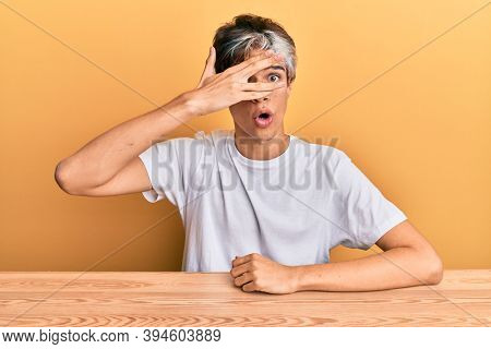 Young hispanic man wearing casual clothes sitting on the table peeking in shock covering face and eyes with hand, looking through fingers afraid 