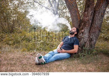 Confident Man Enjoying An Vape On The Grass Of The Forest. Man Really Likes Process Of Smoking.
