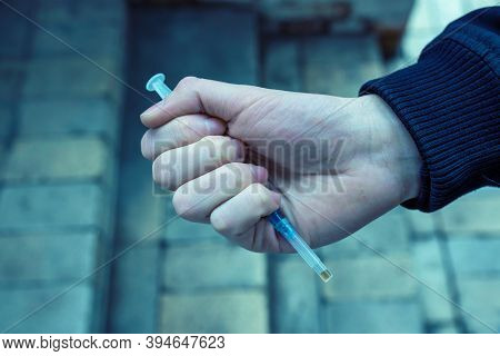Addict Holding A Syringe In His Hand On A Dark Background. Tinted Photo