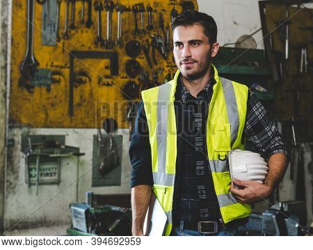 A Young Engineer In Uniform Holding A Hardhat And A Laptop While Standing Within A Metalwork Factory