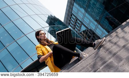 Freelancer With A Laptop Sat On The Steps Near The Business Center, A Young Woman In A Yellow Jacket