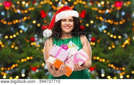 winter holidays, presents and people concept - happy woman in hanta hat holding gift boxes over christmas tree lights background