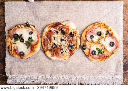 Several Ready-made Mini Pizza, Pizzetta With Sausage And Bacon, Jamon On A Linen Napkin On A Wooden 