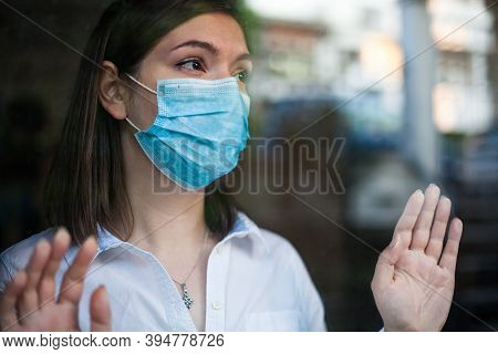 Beautiful Young Woman Wearing Face Mask,looking Through Window Outside,hands Against Glass,coronavir