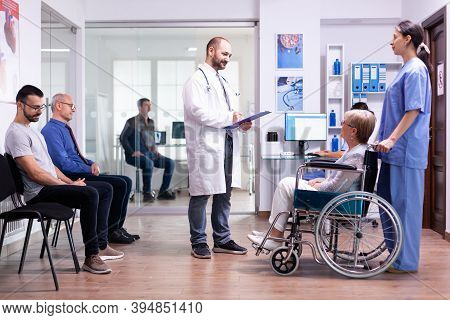 Medic Talking With Disabled Woman In Hospital Waiting Area While Inviting Patient In Examination Roo