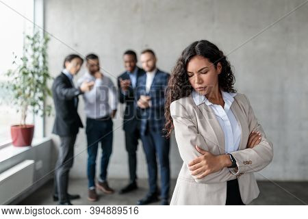Workplace Sexism And Bullying. Unhappy Victimized Businesswoman Standing While Her Male Colleagues W