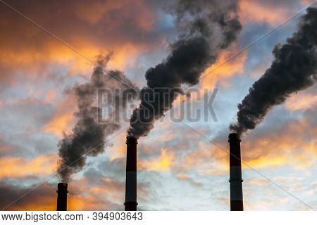 Three Smoking Chimneys In Dramatic Colorful Sky Background. The Concept Of Air Pollution And The Env