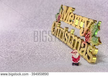 Cute Tiny Figurine Of Santa Claus On A Glitter Silver Snow Background With The Golden Words Merry Ch