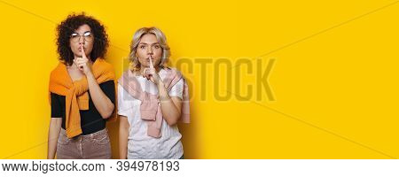 Two Caucasian Women Are Gesturing The Silence Sign On A Yellow Wall With Free Space Looking Seriousl