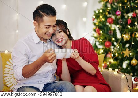 Happy Young Vietnamese Couple Enjoying Hot Chocolate When Resting At Home By Christmas Tree