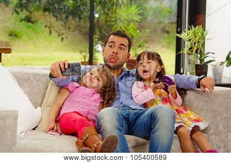 Family portrait of father and two daughters sitting together in sofa posing for selfie making funny