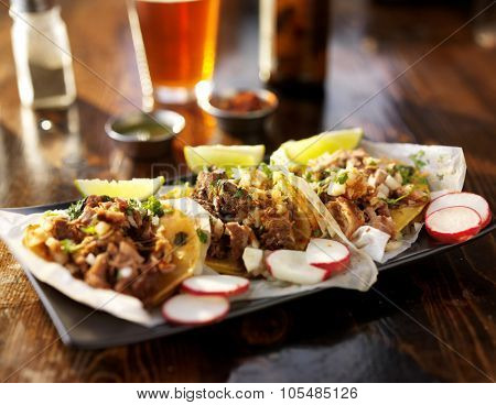 three tacos with beer on wooden table top served with limes, radish slices and beer,  shot with selective focus