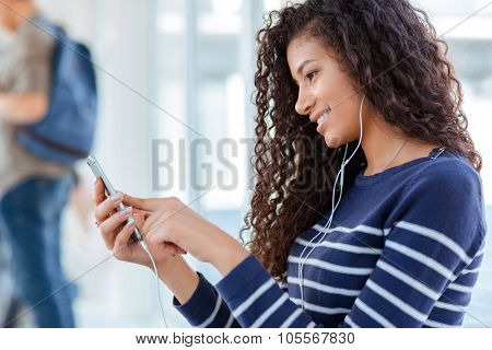 Happy afro american woman using smartphone with headphones in university hall
