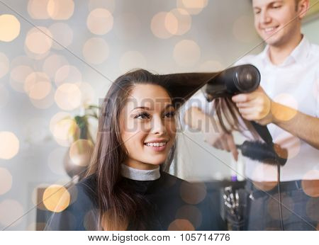 beauty, hairstyle and people concept - happy young woman and hairdresser with fan making hot styling at hair salon over holidays lights