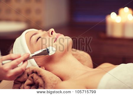 people, beauty, spa, cosmetology and skincare concept - close up of beautiful young woman lying with closed eyes and cosmetologist applying facial mask by brush in spa
