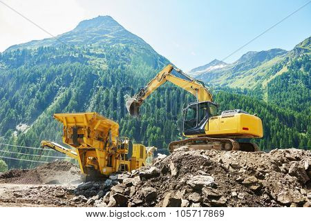 excavator loads the ground in the stone crusher machine during earthmoving works outdoors at mountains construction site