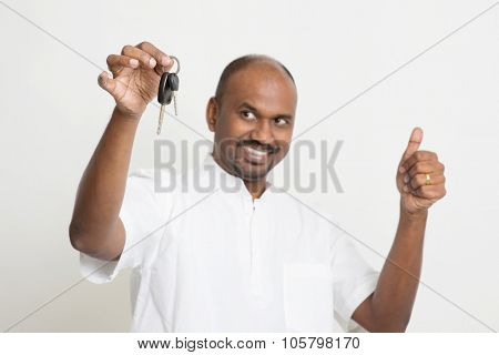 Portrait of Indian man holding new house key and thumb up, real estate property agent concept, focus on key, standing on plain background with shadow.