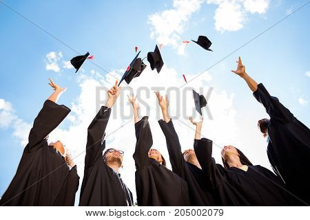 Congratulations to graduates! Low angle shot of cheerful group of six young multi ethnic graduates in black gowns throwing up their hats in the air and celebrating laughing enjoying