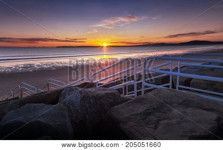 Sunset over Swansea via rock sea defences and a handrail leading to the beach at Aberavon, Port Talbot, UK.