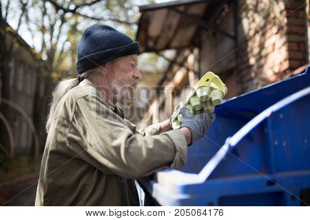 Homeles man searching food in thrash can. View of tramp holding packing for eggs.