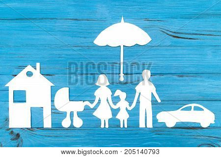 Paper silhouette of family with baby carriage under umbrella, car and house on blue wooden background. Life insurance concept