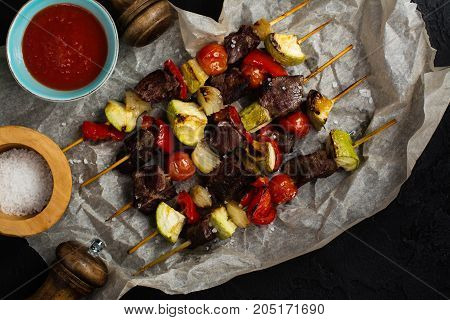 Grilled meat and vegetables cabobs with tomato sauce and sea salt over black stone table. Top view