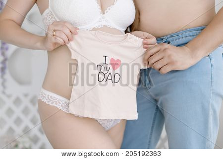 Pregnat woman in tender lingerie and her husband without shirt stand holding pink t-shirt with print I love dad for their unborn baby girl pregnancy concept daylight photoshoot