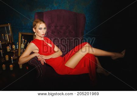 Woman portrait in red dress and high heel shoes. Sitting on stylish violet armchair. Long blond hair with long legs.