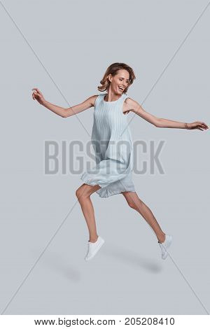 Beauty in the mid-air. Full length of playful young woman gesturing and keeping mouth open while jumping against grey background