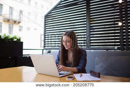 Young woman successful copywriter typing text on portable net-book during rest in restaurant. Beautiful smiling female marketing writer working on laptop computer. Hipster girl using notebook.