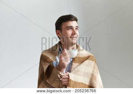 Cozy morning at home. Portrait of hndsome young male with stubble holding white mug having joyful look while relaxing indoors with cup of hot drink warming himself in wool checkered plaid