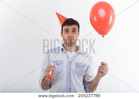 Horizontal portrait of funny upset young Caucasian man wearing holiday cap and blue jeans shirt grimacing feeling unhappy and disappointed that his friends didn't come to his birthday party