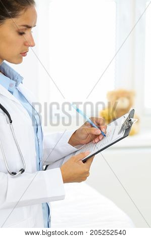 Close-up of a female doctor filling  up medical form at clipboard while standing straight in hospital.