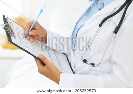 Close-up of a female doctor filling  up medical form at clipboard while standing straight in hospital.