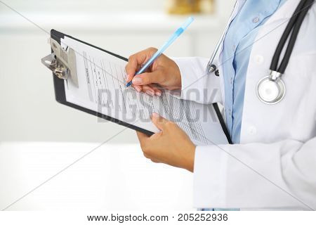 Close-up of a female doctor filling  up medical form at clipboard while standing straight in hospital.