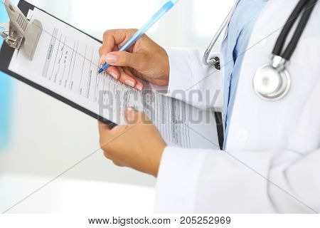 Close-up of a female doctor filling  up medical form at clipboard while standing straight in hospital.
