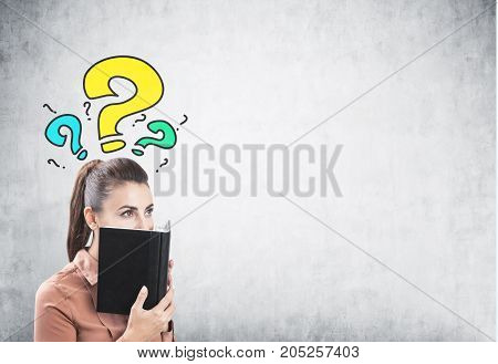 Portrait of a young businesswoman wearing a brown blouse and having long dark hair. She is covering her face with a planner standing near a concrete wall with yellow and blue question marks. Mock up