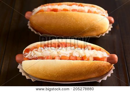 Chilean Completo Clasico (classical) or Completo Aleman (German) traditional hot dog sandwiches made of bread sausage tomato cubes and sauerkraut photographed on dark wood with natural light (Selective Focus Focus in the middle of the first hotdog)