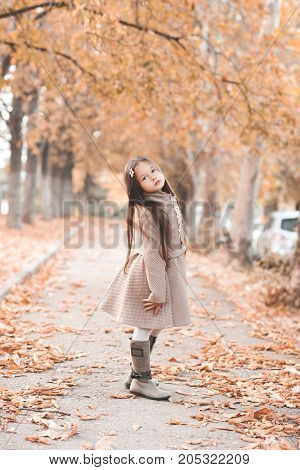 Stylish kid girl 4-5 year old wearing trendy winter jacket and boots standing in street aith fallen leaves. Looking at camera. Autumn season.