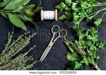 Bouquet garni. Classical french composition. Bay leaf parsley and thyme bound with culinary thread. Bunch of fresh aromatic herbs coil with threads old scissors on black textured background