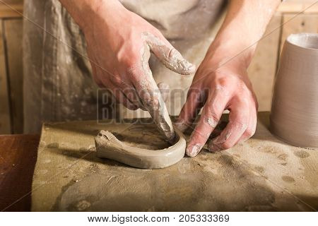 pottery, stoneware, ceramics art concept - closeup on craftsman fingers forming the handle from raw fireclay, master hands work with some piece of clay, male stand at a workshop near the table