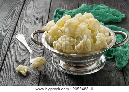 Blanched cauliflower in colander on gray wooden background. White cauliflower inflorescences in metal colander fork and green gauze napkin on table. Preparing for diet dinner