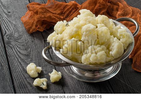 Blanched cauliflower in colander on gray wooden background. White cauliflower inflorescences in metal colander brown gauze napkin on table. Preparing for diet dinner