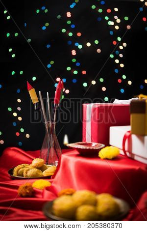 stock photo showing Diwali still life with Diya or oil Lamp, gift boxes, sweets and fire crackers with flowers arranged over red satin cloth with bokeh
