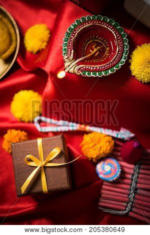 stock photo showing Diwali still life with Diya or oil Lamp, gift boxes, sweets and fire crackers with flowers arranged over red satin cloth with bokeh