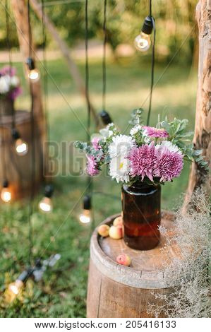 cosy atmosphere, happiness, nature concept. composition made of lovely bunch of purple and white asters in the botel of dark brown glass standing on the wooden barrel nearby twinkle lights